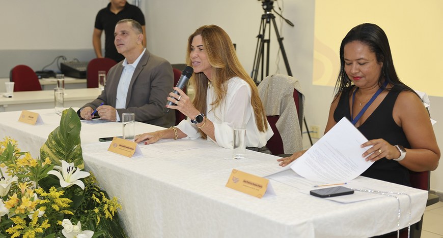 Mesa de evento com três pessoas sentadas atrás de uma toalha branca. Ao centro, uma mulher de ca...