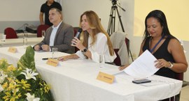 Mesa de evento com três pessoas sentadas atrás de uma toalha branca. Ao centro, uma mulher de ca...