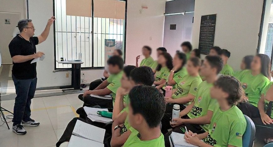 Em uma sala de aula clara, um homem em pé, à esquerda, conduz uma atividade enquanto aponta para...