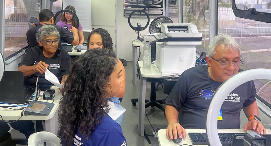 Em uma sala de trabalho, várias pessoas estão sentadas em estações com computadores. Em primeiro...