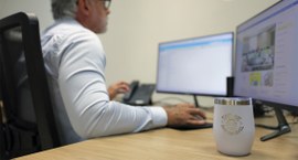 Homem de cabelos grisalhos e óculos trabalha sentado diante de dois monitores em uma mesa de esc...